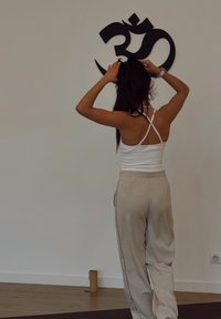 Débardeur blanc à fines bretelles et pantalon de survêtement beige avec boutons latéraux, debout devant un symbole 'Om' noir sur un mur blanc.