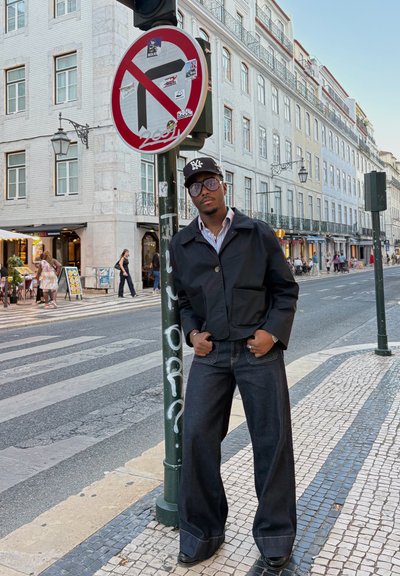 Chaqueta negra recortada, camisa a rayas y jeans anchos de mezclilla oscura. Calle de la ciudad, señal de tráfico y edificios al fondo.