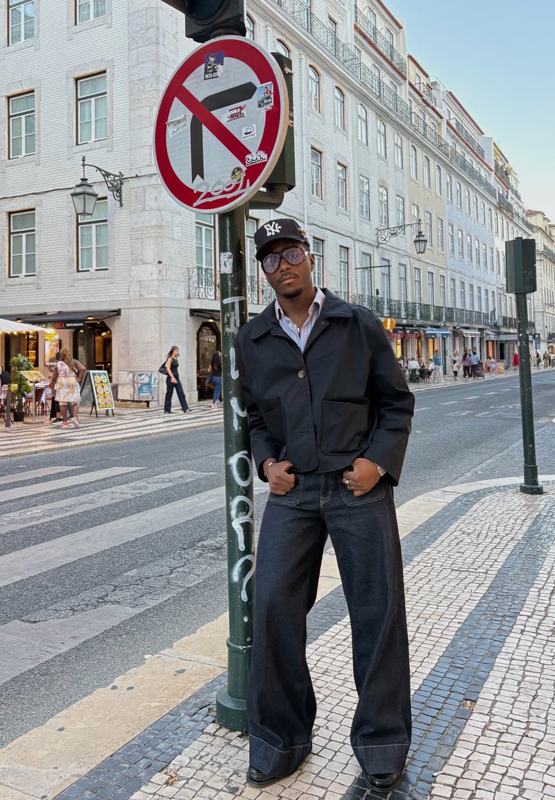 Veste noire coupée, chemise rayée et jean en denim foncé à jambes larges. Rue de la ville, panneau de signalisation et bâtiments en arrière-plan.