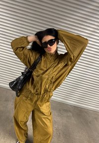 Khaki kombinezon z zapinaniem na zamek z przodu, długimi rękawami i elastycznymi mankietami. Czarna torba na ramię z detalami w paski i dużymi okularami przeciwsłonecznymi. Teksturowane wykończenie.