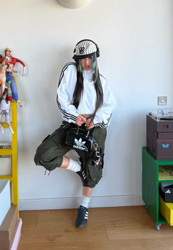 Mladá osoba v bielej bunde Adidas, šiltovke a cargo nohaviciach pózuje v interiéri, drží čiernu tašku Adidas, popri ktorej je farebné bábiky na žltej polici.