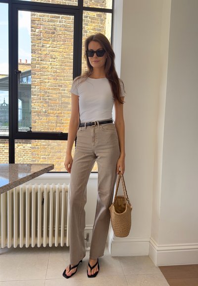 Mujer con gafas de sol, camiseta blanca, pantalones beige, cinturón negro y sandalias, sosteniendo un bolso tejido en el interior cerca de una ventana y un radiador.