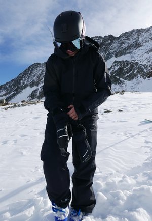 Chaqueta y pantalones de esquí impermeables en negro, con guantes negros, usados en terreno nevado. Se ven botas de esquí azules. Montañas en el fondo.