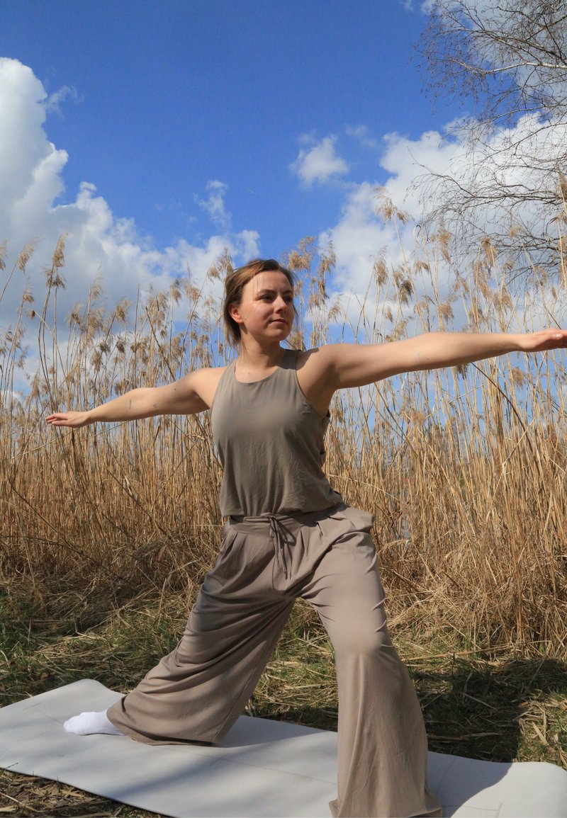 Femme en vêtements beige amples pratique une posture de yoga sur un tapis en extérieur près de roseaux secs sous un ciel bleu.