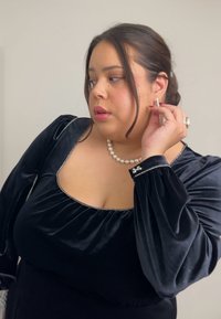 Bluza din catifea neagră cu mâneci bufante, decolteu pătrat și accente decorative argintii. Poartă un colier de perle și bijuterii argintii.