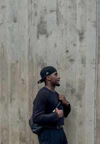 Maglione nero a maniche lunghe, pantaloni scuri e un cappellino. Tiene in mano una borsa nera. Sfondo in texture legno, colori neutri. Design casual.