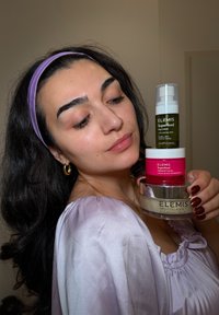 Tre Elemis hudvårdsprodukter: en grön mist, en rosa kräm och en beige rengöringsbalm, mot en neutral bakgrund, hållna av en person.