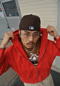 Sweatshirt vermelho com fecho à frente e gola alta, usado sobre uma t-shirt gráfica castanha. Os acessórios incluem correntes prateadas e óculos redondos, inseridos em frente a uma parede de tijolo.
