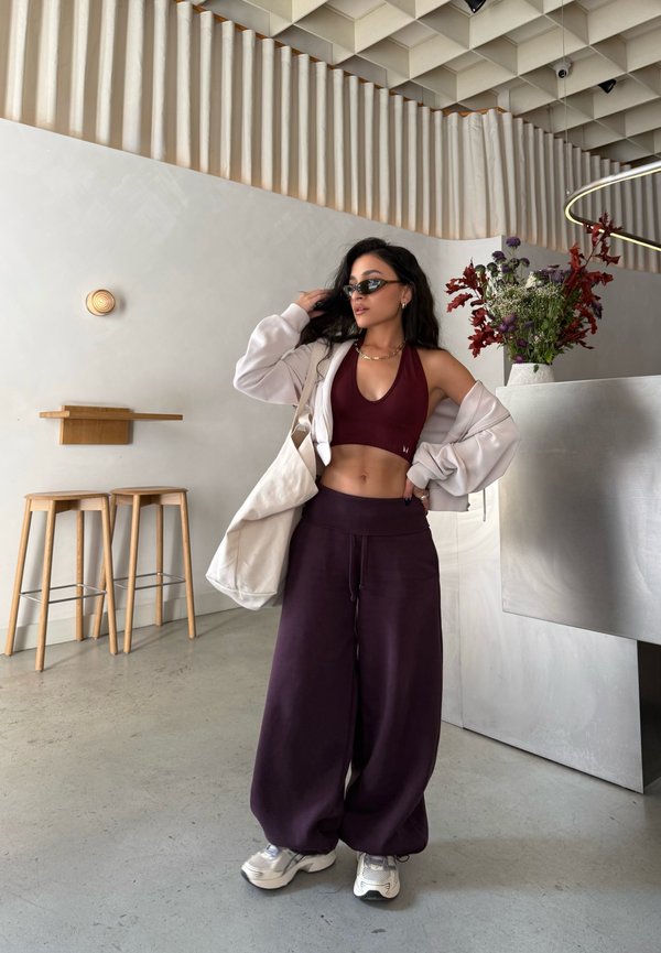 Mujer con top halter burdeos y pantalones, con chaqueta beige y bolso tote, de pie en una cafetería moderna y minimalista con taburetes y flores.