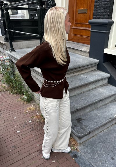 Cárdigan marrón con cierre de botones, combinado con pantalones de pierna ancha color crema que cuentan con botones laterales. Los accesorios incluyen un cinturón de cuentas.