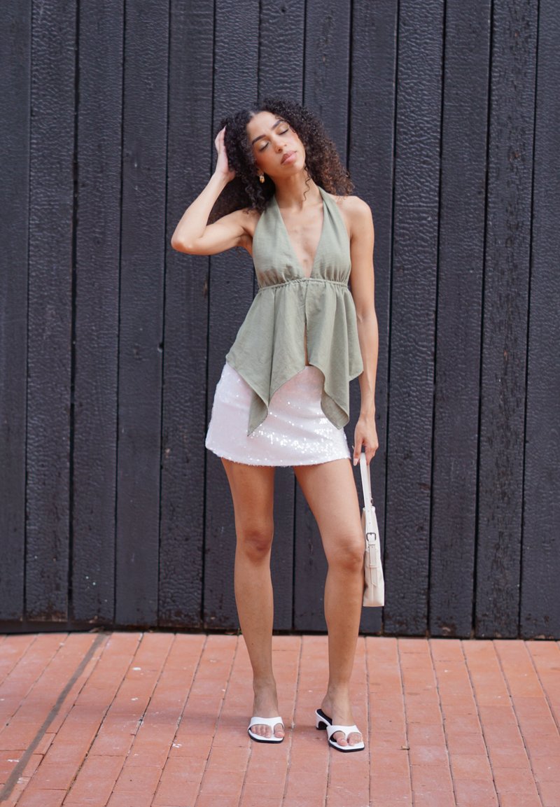 Femme aux cheveux bouclés portant un haut sans manches vert, une jupe blanche à sequins, des sandales blanches, tenant un petit sac à main beige contre un mur en bois foncé.