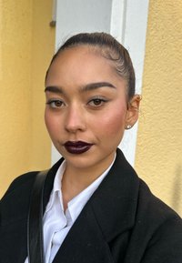 Portret van een persoon met recht, donker haar in een knot, gekleed in een zwarte jas en een wit hemd, met donkere lippenstift en natuurlijke make-up.