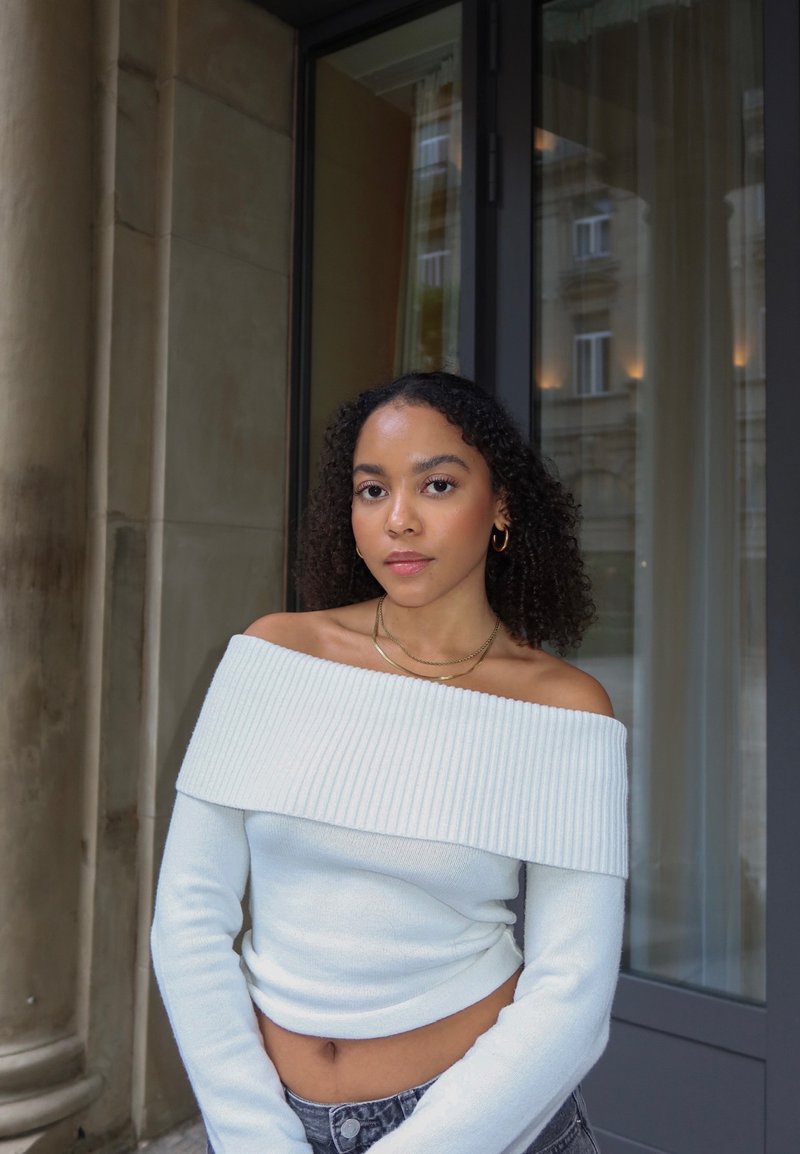 Pull blanc à épaules dénudées avec une texture côtelée, manches longues et coupe courte, associé à un jean en denim gris et des colliers en or superposés.