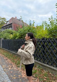 Donna con giacca beige e pantaloni neri che sistema il colletto accanto a una recinzione di legno nera, con arbusti verdi e una casa con tetto rosso sullo sfondo.