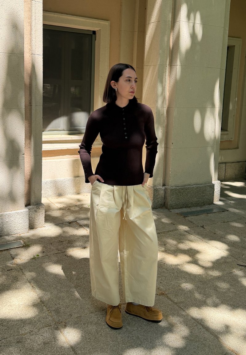 Femme debout à l'extérieur sous une lumière mouchetée, portant un haut foncé ajusté, un pantalon crème ample et des chaussures brunes, les mains dans les poches, tournée vers la gauche.