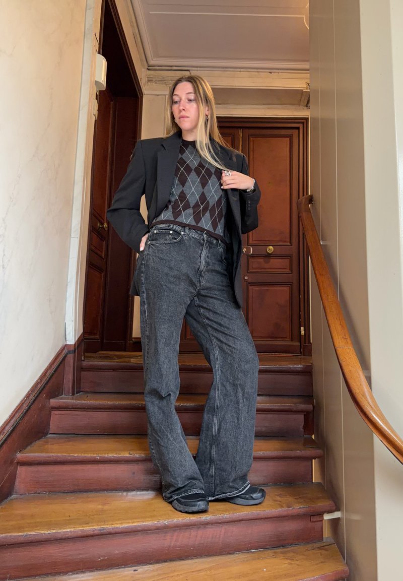 Vrouw staat op houten trappen, draagt een zwart blazer, een argyle trui, losse grijze jeans en zwarte schoenen, binnen bij een houten deur.