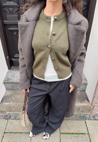 Oversized grijze wollen jas over een groene gebuttonde cardigan en een wit hemd. Losse zwarte broek en zwart-wit patroon schoenen met een handtas.