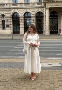 Ženska v beli čipkasti majici in dolgi krilni nosi očala, stoji na tlakovanem pločniku blizu prehoda za pešce in zgodovinske fasade stavbe.
