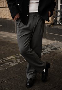 Grå ullbukser med avslappet passform, sidelommer og smalnet ben. Kombinert med polerte svarte loafers og en svart jakke.