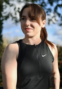 Débardeur de sport noir avec une fermeture éclair et des accents en mesh, arborant un petit logo Nike blanc sur la poitrine. L'arrière-plan montre un feuillage flou.