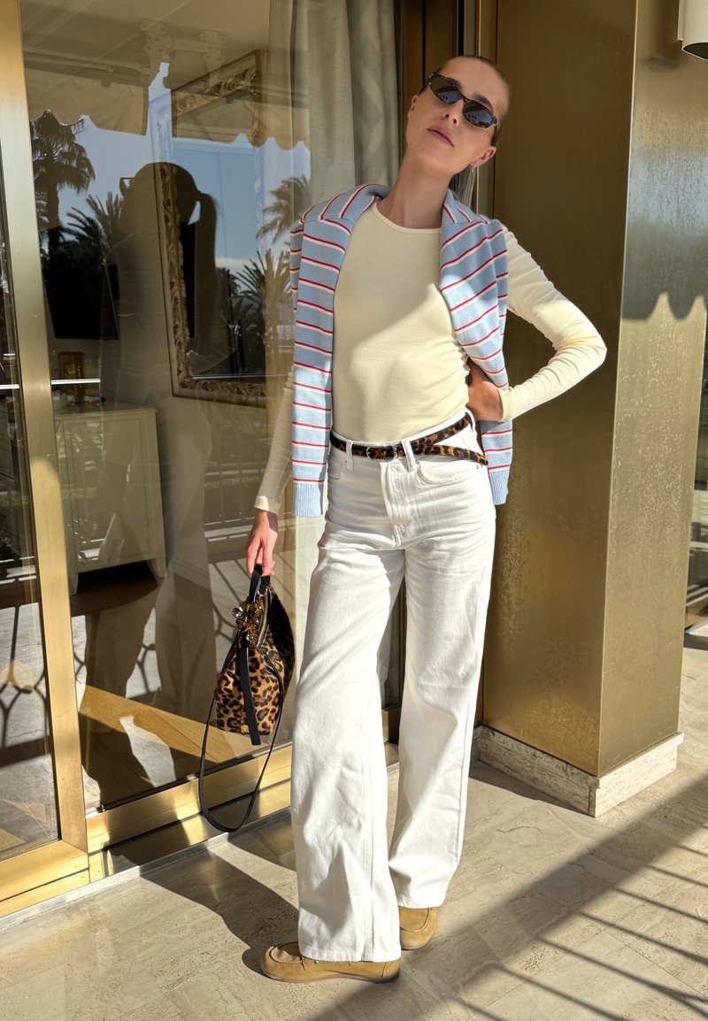 Femme portant des lunettes de soleil, haut crème à manches longues, pull rayé sur les épaules, pantalon blanc à jambes larges, chaussures beige, tenant un sac à motif léopard.