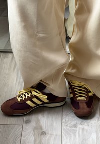 Tênis em marrom e castanho com detalhes amarelos, apresentando três riscas, materiais em camurça e rede, e atacadores redondos, combinados com umas calças creme.