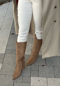 Beige Wildleder-Stiefel in kniehoher Form mit spitzem Fuß und Blockabsatz, ausgestattet mit einem seitlichen Reißverschluss und einer weichen Textur, kombiniert mit weißen engen Hosen.