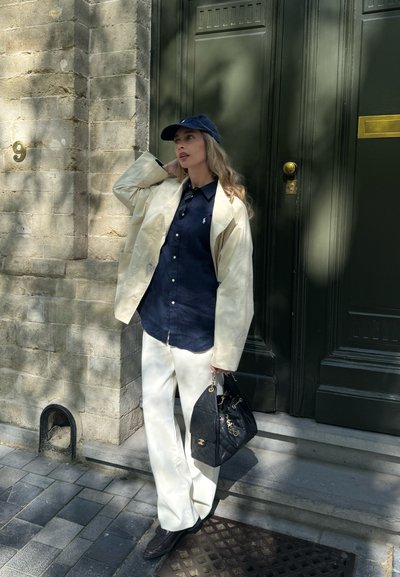 Mujer con blazer beige, camisa azul marino, pantalones blancos y gorra azul marino, de pie fuera de un edificio de piedra sosteniendo un bolso negro.