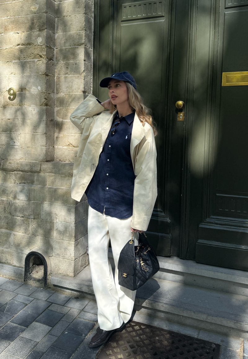Γυναίκα με μπεζ σακάκι, μπλε πουκάμισο, άσπρο παντελόνι και μπλε καπέλο στέκεται έξω από πέτρινο κτίριο κρατώντας μαύρη τσάντα.