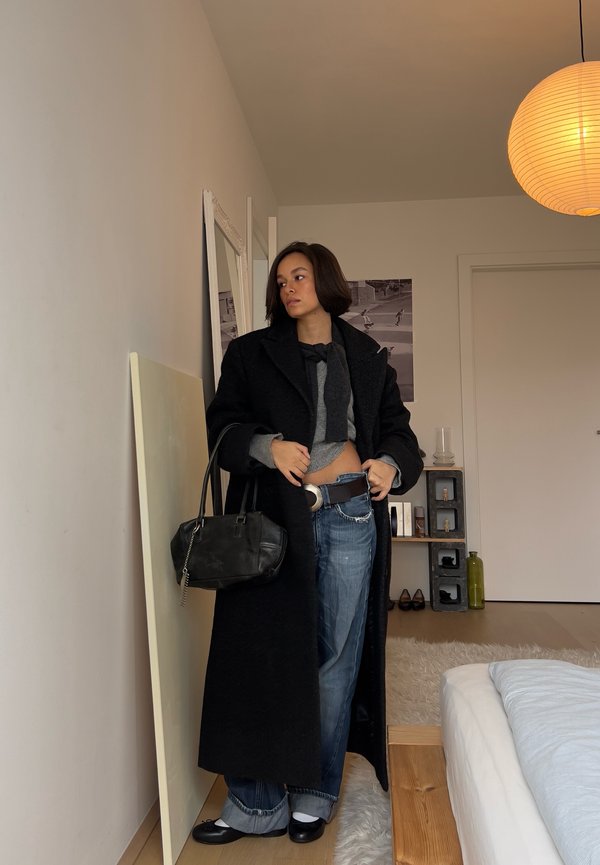 Žena stojící v místnosti, má na sobě dlouhý černý kabát, šedý svetr, volné džíny, černé boty, drží černou kabelku a dívá se stranou.