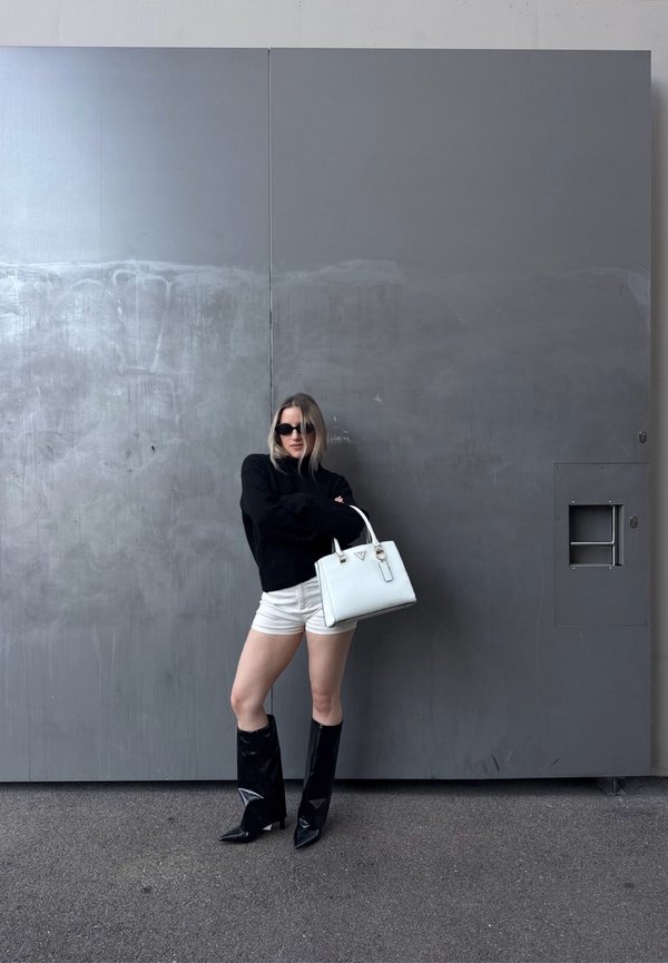 Mujer con suéter negro, pantalones cortos blancos, botas negras, gafas de sol, sosteniendo un bolso blanco, de pie contra una pared industrial gris al aire libre.