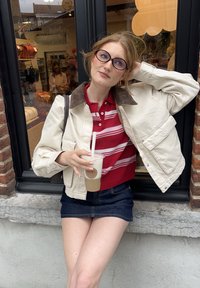 Kremfarget jakke med brun kordfløyelskrage, rød og hvit stripet polo, mørke denimshorts, holder en drink, står mot et vindu.