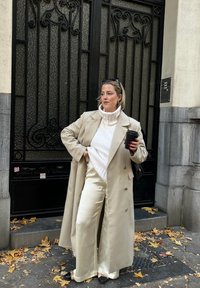 Femme en trench-coat beige, col roulé crème et pantalon en satin tenant une tasse noire, debout sur un trottoir couvert de feuilles devant une grille en fer noir.