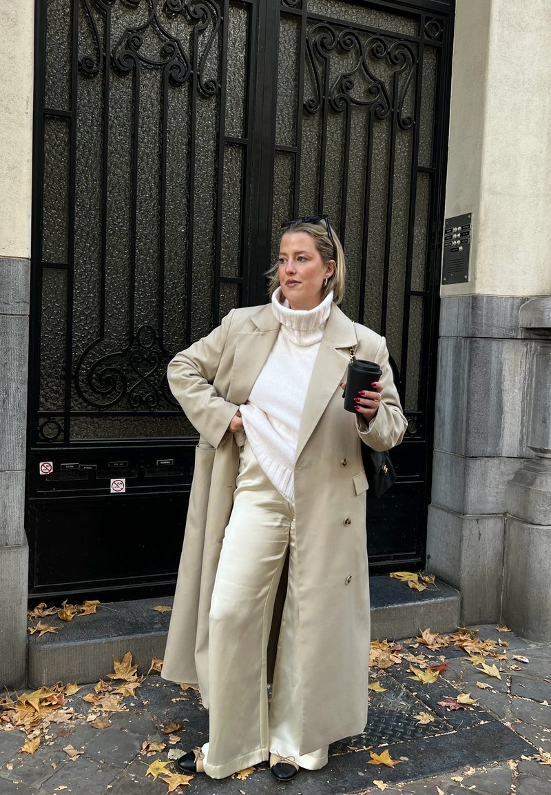 Femme en trench-coat beige, col roulé crème et pantalon en satin tenant une tasse noire, debout sur un trottoir couvert de feuilles devant une grille en fer noir.