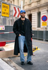 Cappotto nero, maglione nero, jeans azzurri chiari e scarpe nere. Cappellino da baseball blu con logo. Ambiente urbano con cartelli di costruzione.