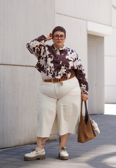 Camisa: recortada, estampado animal en marrón y blanco; pantalones: de pierna ancha en beige claro; zapatos: de suela gruesa en beige con cordones marrones; bolso marrón; tatuajes visibles.