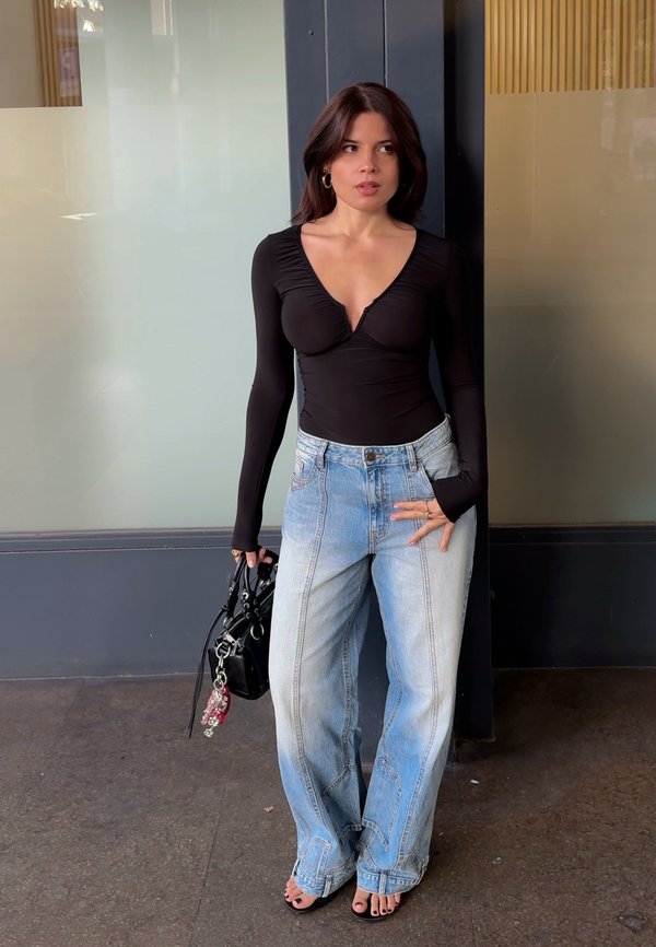 Une femme portant un haut noir et un jean ample se tient près d’une vitrine en verre, tient un sac à main noir et regarde sur le côté.