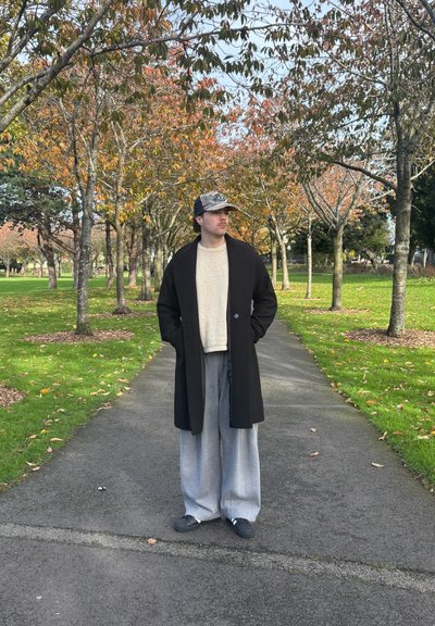 Abrigo largo negro, suéter beige claro, pantalones anchos grises, zapatos negros y gorra gris. Al aire libre con árboles y hojas de otoño en el fondo.