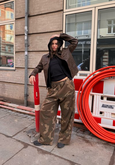 Chaqueta de cuero marrón con capucha, top negro corto, pantalones de camuflaje de pierna ancha y zapatos negros; se encuentra al lado de una barrera de tráfico roja.