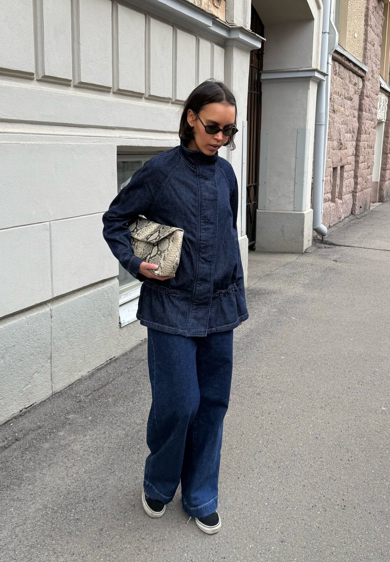 Femme portant une veste et un pantalon en denim foncé, tenant une pochette en peau de serpent, marchant sur un trottoir en ville près de bâtiments en pierre.