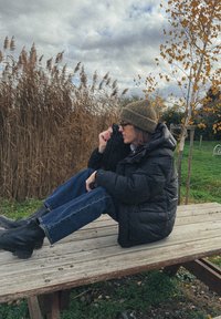 Γυναίκα με μαύρο μπουφάν, πράσινο σκουφάκι και μπότες κάθεται σε ξύλινο τραπέζι πικνίκ, με ψηλό ξερό γρασίδι και φθινοπωρινούς δέντρους στο βάθος.