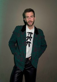 Άνδρας με κοντά μαλλιά και γένια, ντυμένος με πράσινο καρό σακάκι, λευκό πουκάμισο με μαύρα φιόγκους από κορδέλα και μαύρο δερμάτινο παντελόνι, που στέκεται μπροστά σε έναν απλό τοίχο.