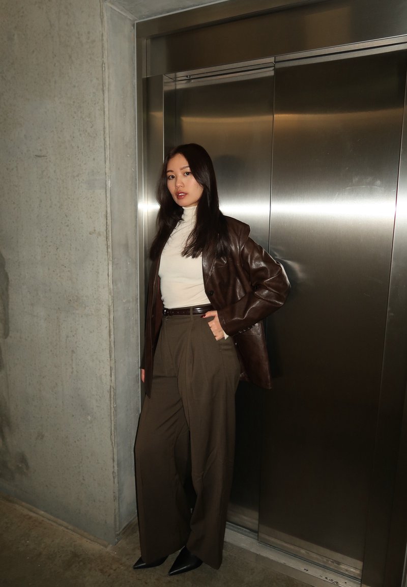 Femme portant une veste en cuir marron, un col roulé blanc et un pantalon large, debout avec une main dans la poche près d'une porte d'ascenseur fermée.