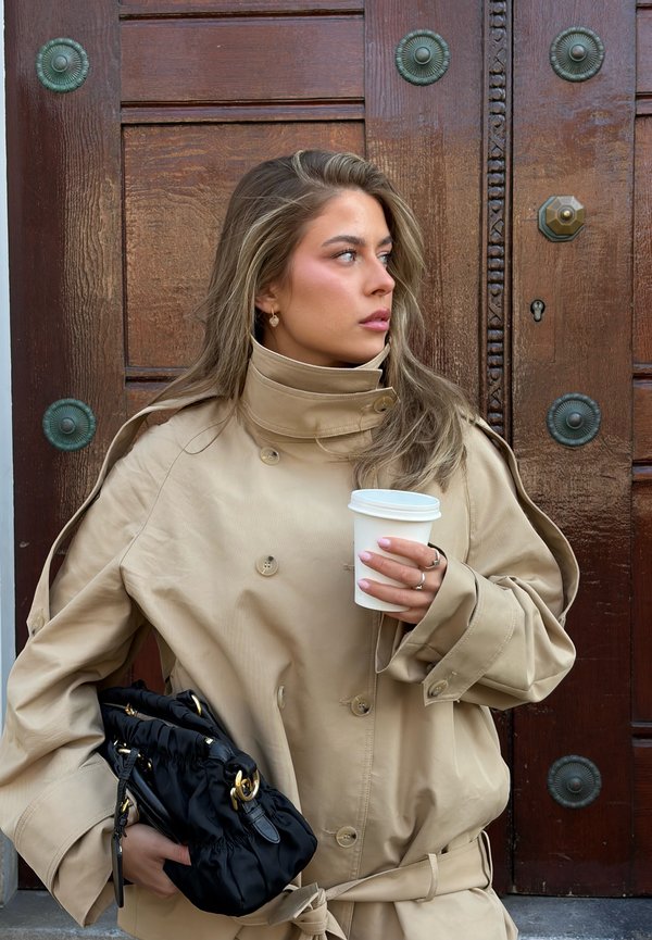 Młoda kobieta w beżowym trenczu trzymająca filiżankę kawy i czarną torebkę, stojąca na tle szczegółowych drewnianych drzwi.