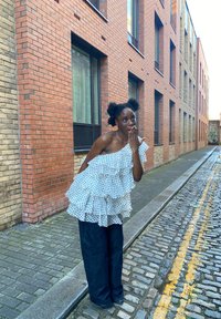 Fehér pöttyös blúz fodros rétegekkel, sötétkék, bő szárú nadrággal párosítva. Az utcakép macskakövekkel és téglából készült épületekkel tarkított.