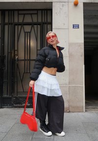 Personne aux cheveux courts roses portant des lunettes de soleil rouges, une doudoune courte noire, une jupe blanche superposée sur un pantalon noir large, tenant un sac rouge.