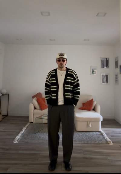Hombre con cárdigan de rayas, camisa blanca, pantalones oscuros, gafas de sol y gorra beige de pie en una sala de estar con sofá blanco y arte enmarcado en la pared.