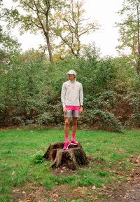 Athlete standing on a tree stump, dressed in a light grey long-sleeve shirt with pink accents, grey shorts, pink socks, and pink trainers. Background of green foliage.
