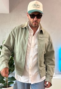 Heledad roheline lukuga jakk kraega, mille all on valge nööbitav särk ja hallid teksad. Aksessuaarideks päikeseprillid ja beež nokamüts.