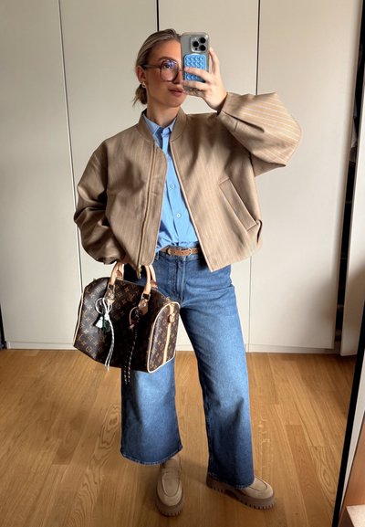 Chaqueta bomber a rayas marrón, camisa de botones azul claro, vaqueros de pierna ancha, mocasines beige de plataforma y bolsos marrón de Louis Vuitton con accesorios.